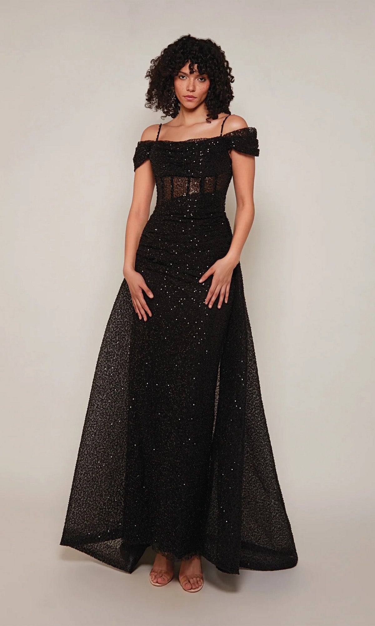 A person stands against a plain background wearing the Alyce 7101—a long, black formal dress with thin straps, off-the-shoulder style, and sparkling glitter details.
